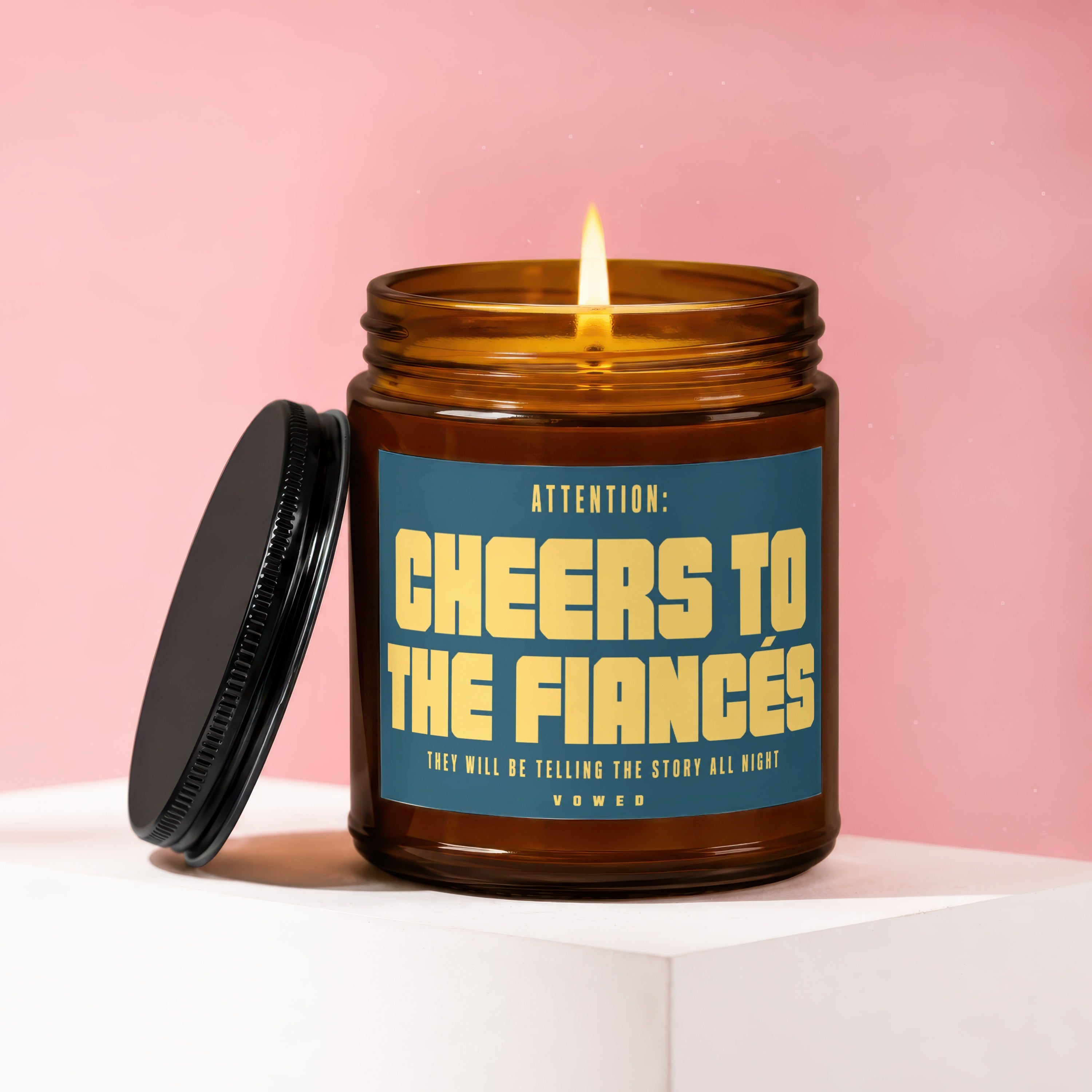 Amber glass candle with lit flame, lid ajar, reads "Cheers to the Fiancés".