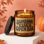 Amber glass candle with lit flame and "Smells Like Someone Else's Open Bar" label.