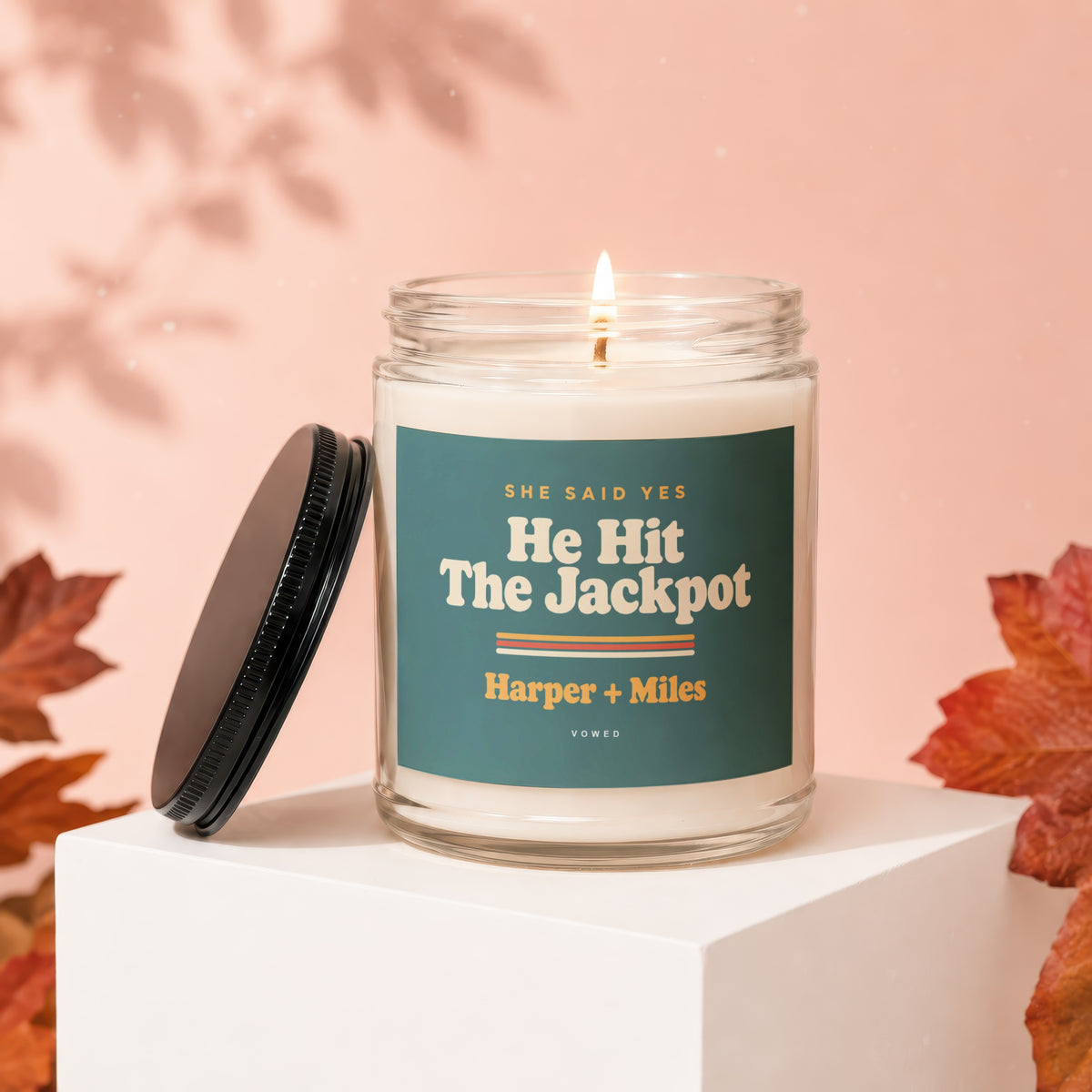Candle with label "She Said Yes He Hit The Jackpot Harper + Miles" on a white cube.