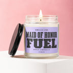 Glass jar candle with "Maid of Honor Fuel" label and lit wick, pink background.