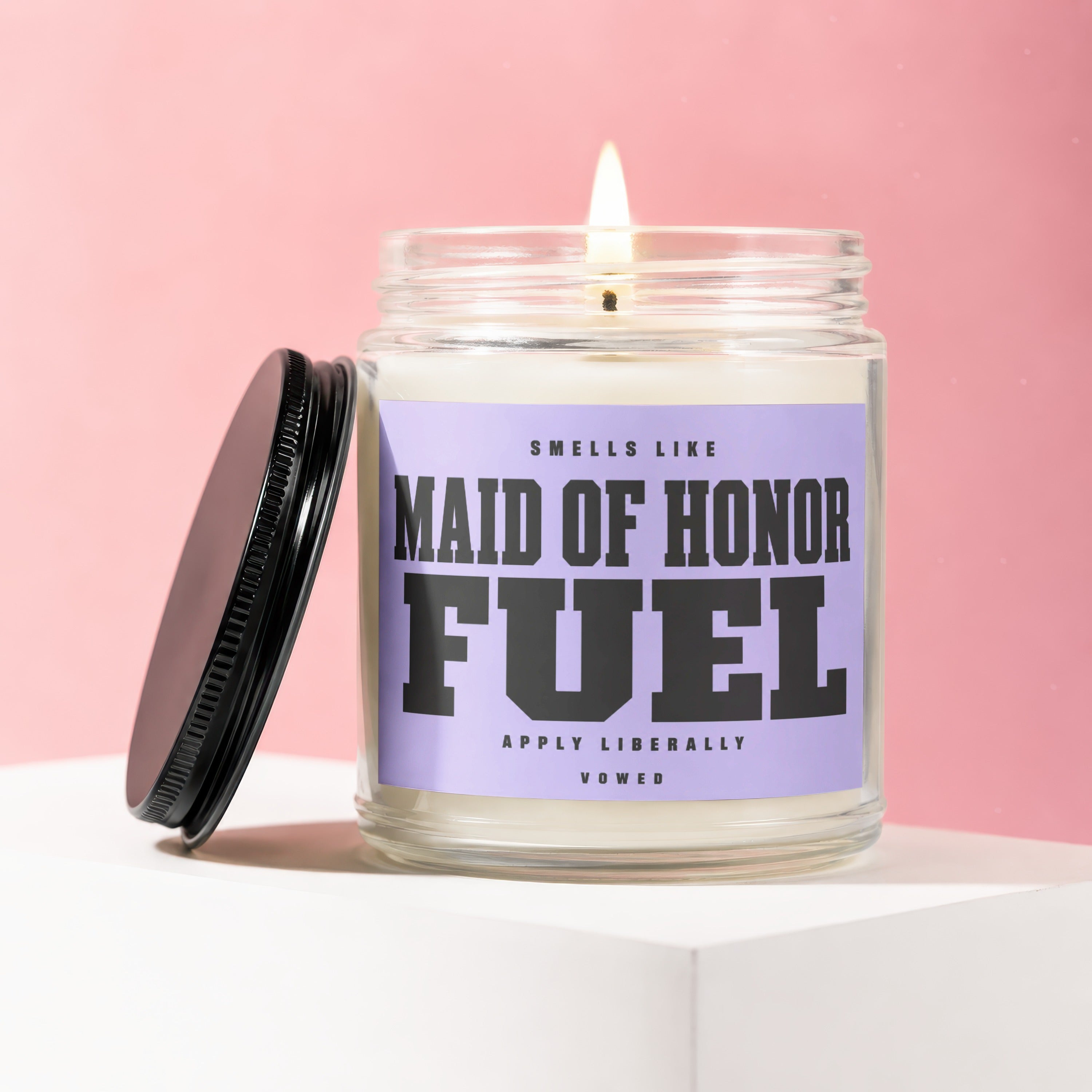 Glass jar candle with "Maid of Honor Fuel" label and lit wick, pink background.