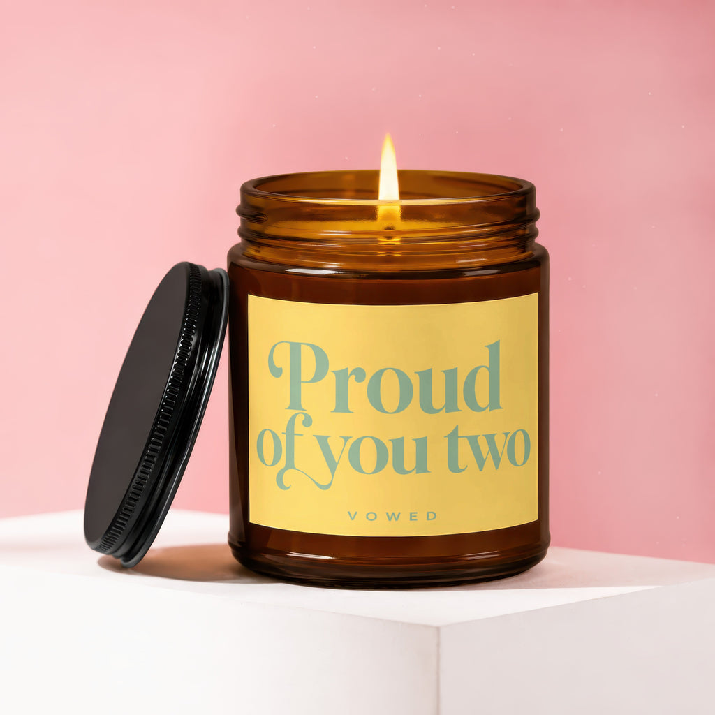 Lit amber glass candle with yellow label reading "Proud of you two, VOWED".