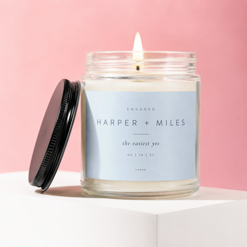 Glass candle with lit wick, featuring label "Engaged Harper + Miles the easiest yes 06 | 14 | 27"