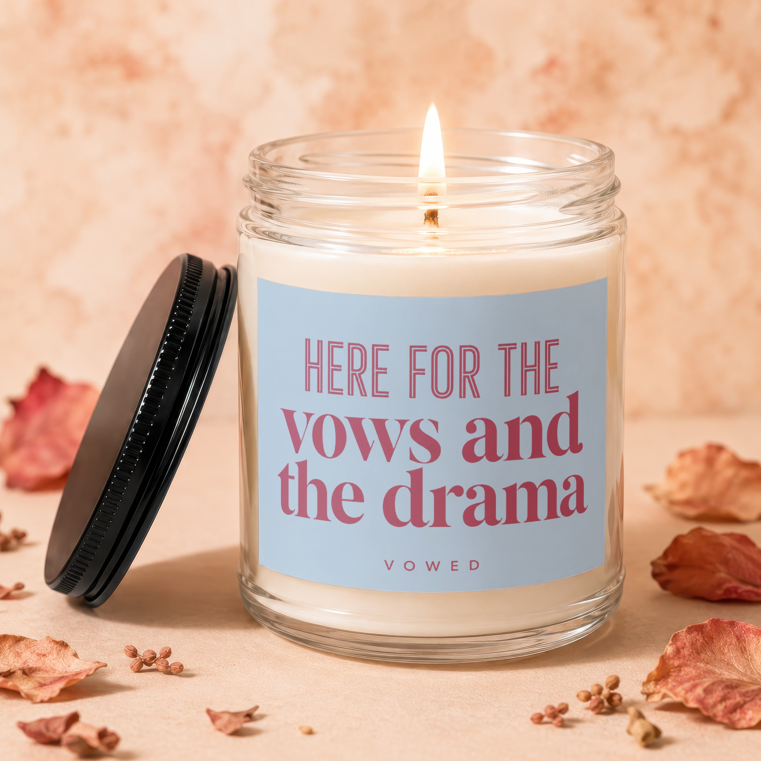 Lit candle in a glass jar with a blue label reading "HERE FOR THE vows and the drama"