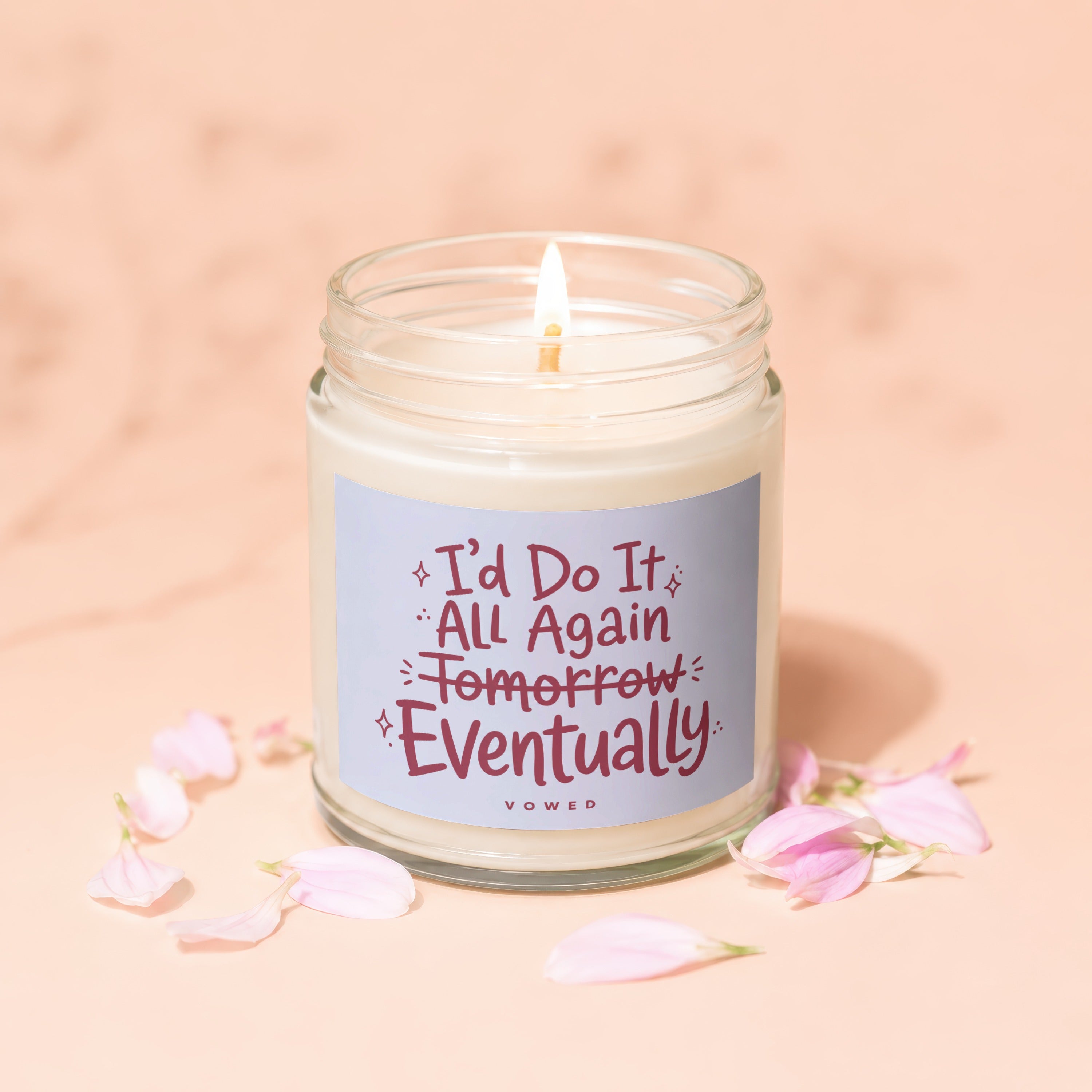Glass jar candle with burning wick, surrounded by pink petals, reads "I'd Do It All Again Tomorrow Eventually".