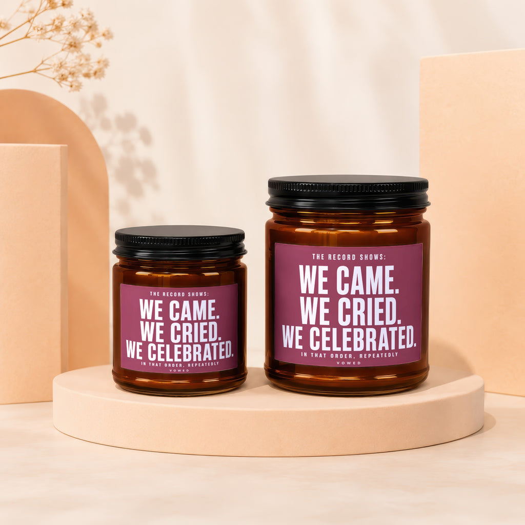 Two amber glass candles with black lids and purple labels reading "WE CAME. WE CRIED. WE CELEBRATED."