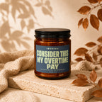 Amber jar candle with a blue label reading "INVOICE: CONSIDER THIS MY OVERTIME PAY".
