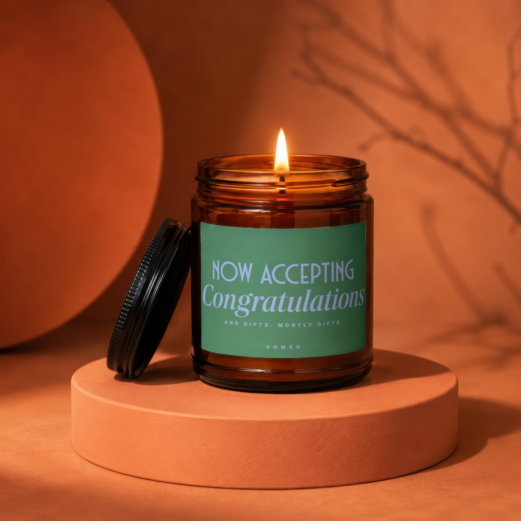 Lit amber glass candle with green label reading "NOW ACCEPTING Congratulations AND GIFTS. MOSTLY GIFTS. VOWED."