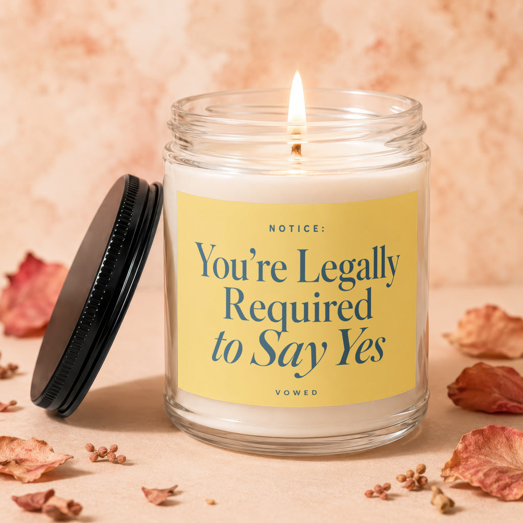 Glass candle with a lit wick and a yellow label reading "NOTICE: You're Legally Required to Say Yes".