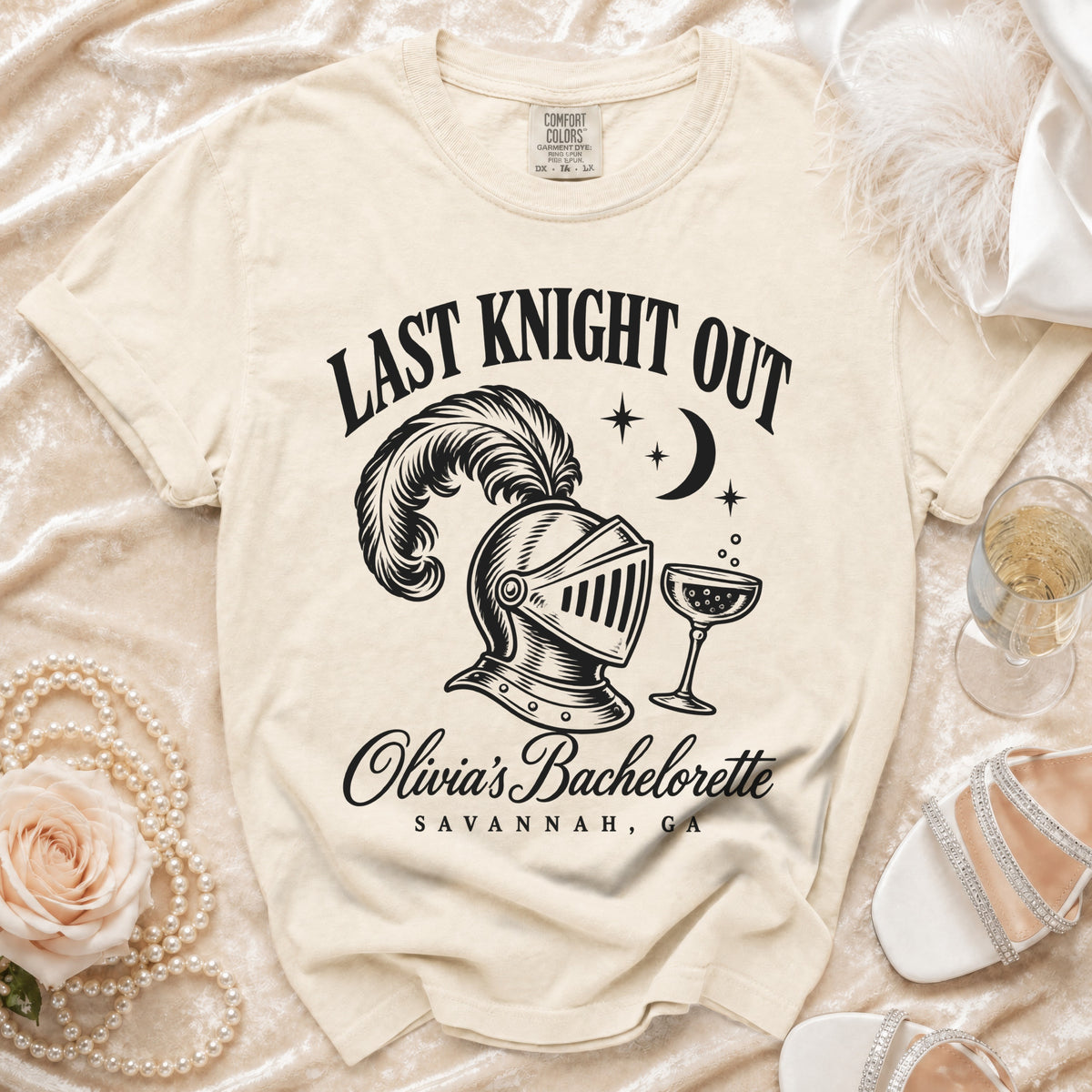 Cream t-shirt featuring a black graphic with a plumed knight helmet, champagne glass, and text reading LAST KNIGHT OUT OLIVIA'S BACHELORETTE SAVANNAH GA.