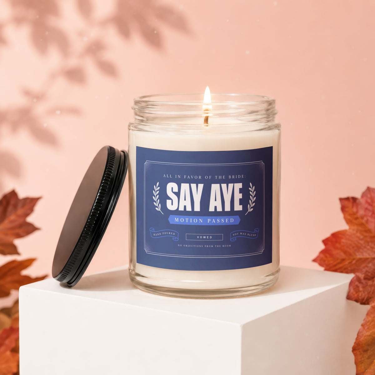 Glass candle with "Say Aye" label, lid off, near autumn leaves.
