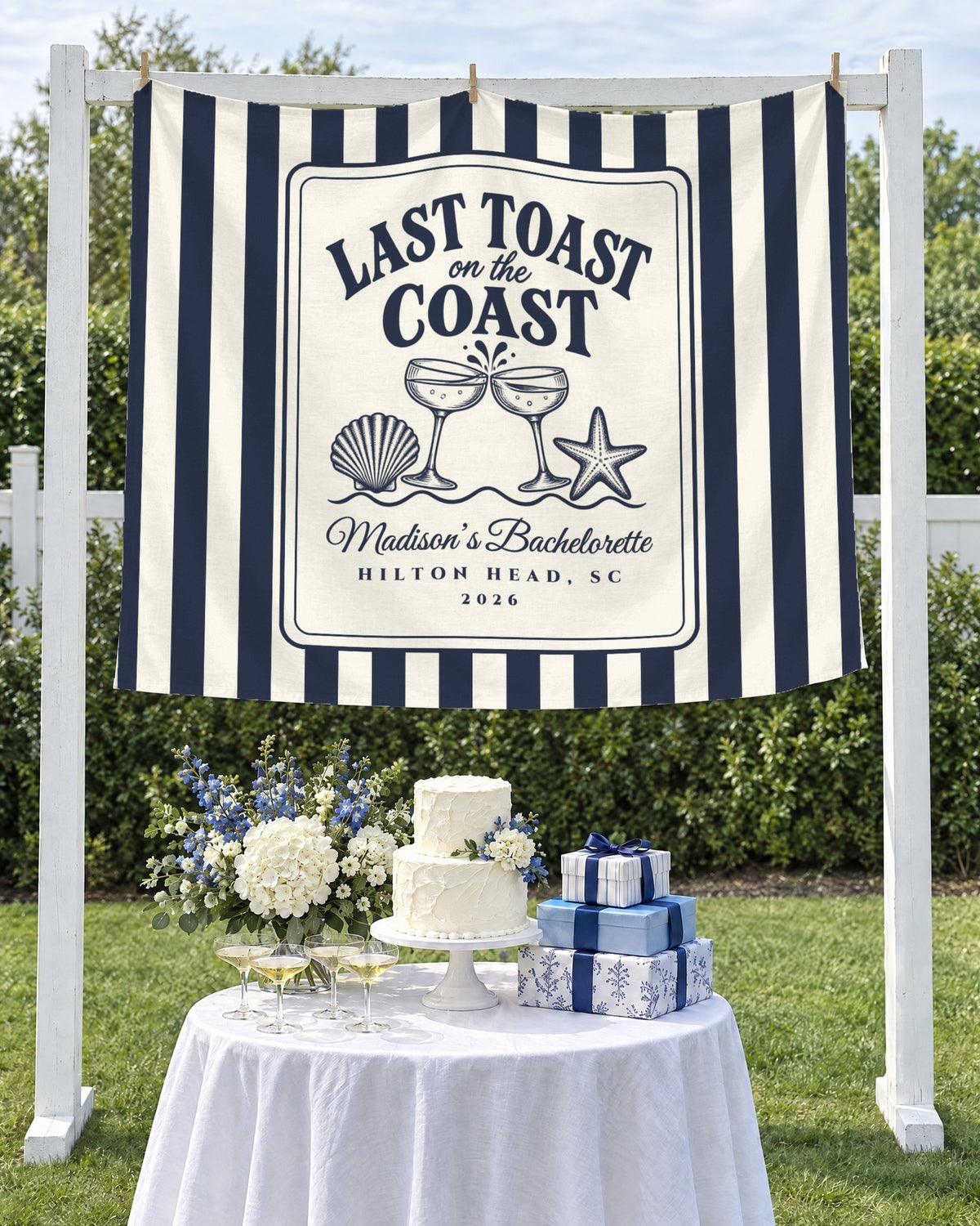 Last Toast On The Coast striped bachelorette tapestry hung as a backdrop over a cake table with champagne coupes in a garden
