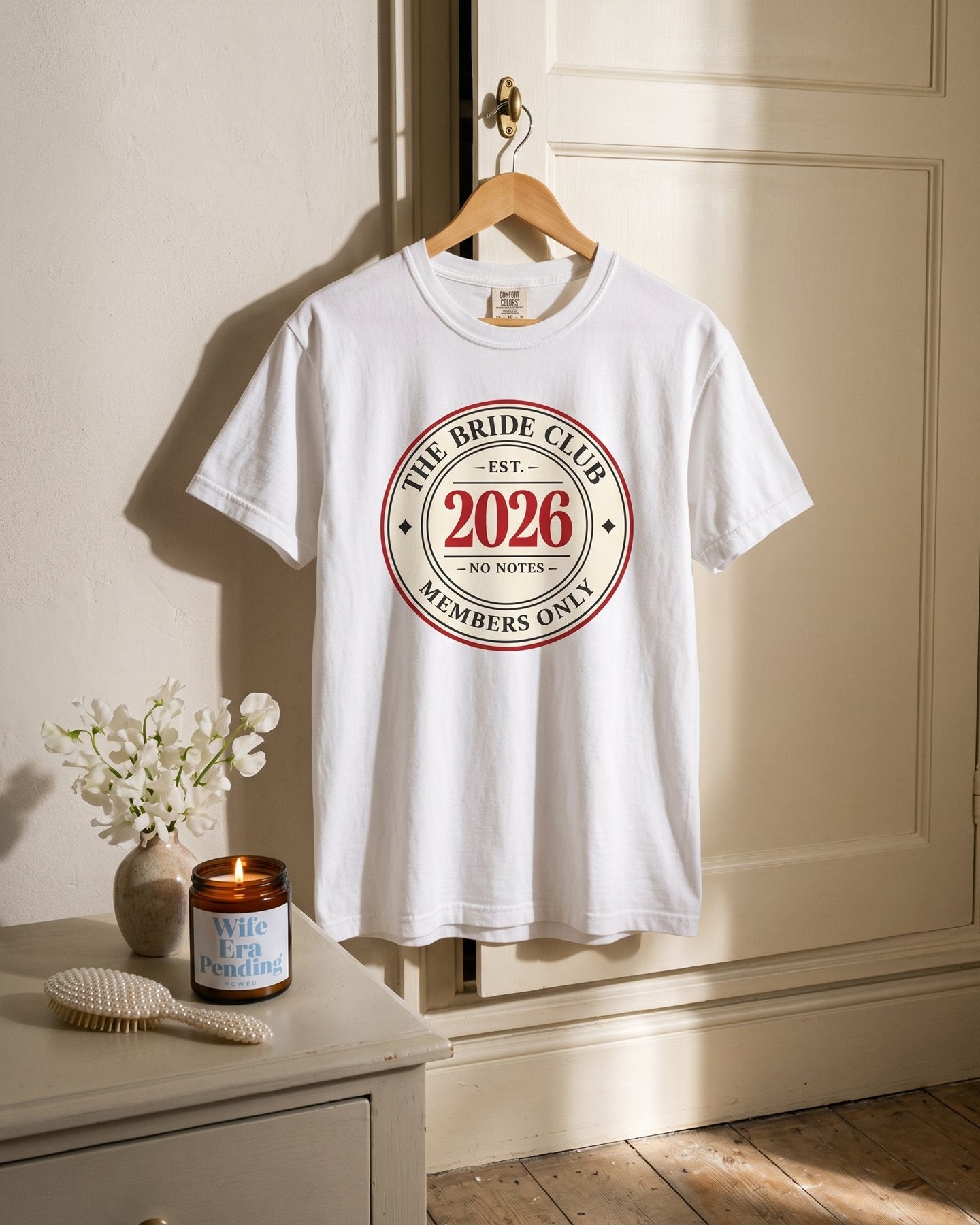 The Bride Club 2026 tee on a wooden hanger above a lit Wife Era Pending candle in a sunny villa bedroom