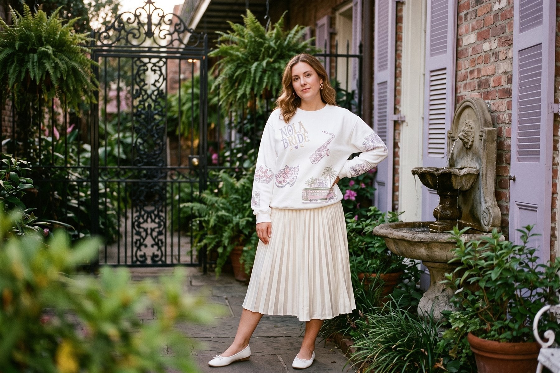 Bride-to-be in the NOLA Bride doodles sweatshirt and a pleated ivory skirt in a French Quarter courtyard